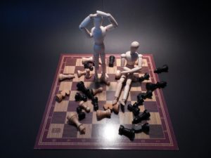 Two small wooden drawing dummies standing on a chess board with chess pieces knocked down everywhere around them.