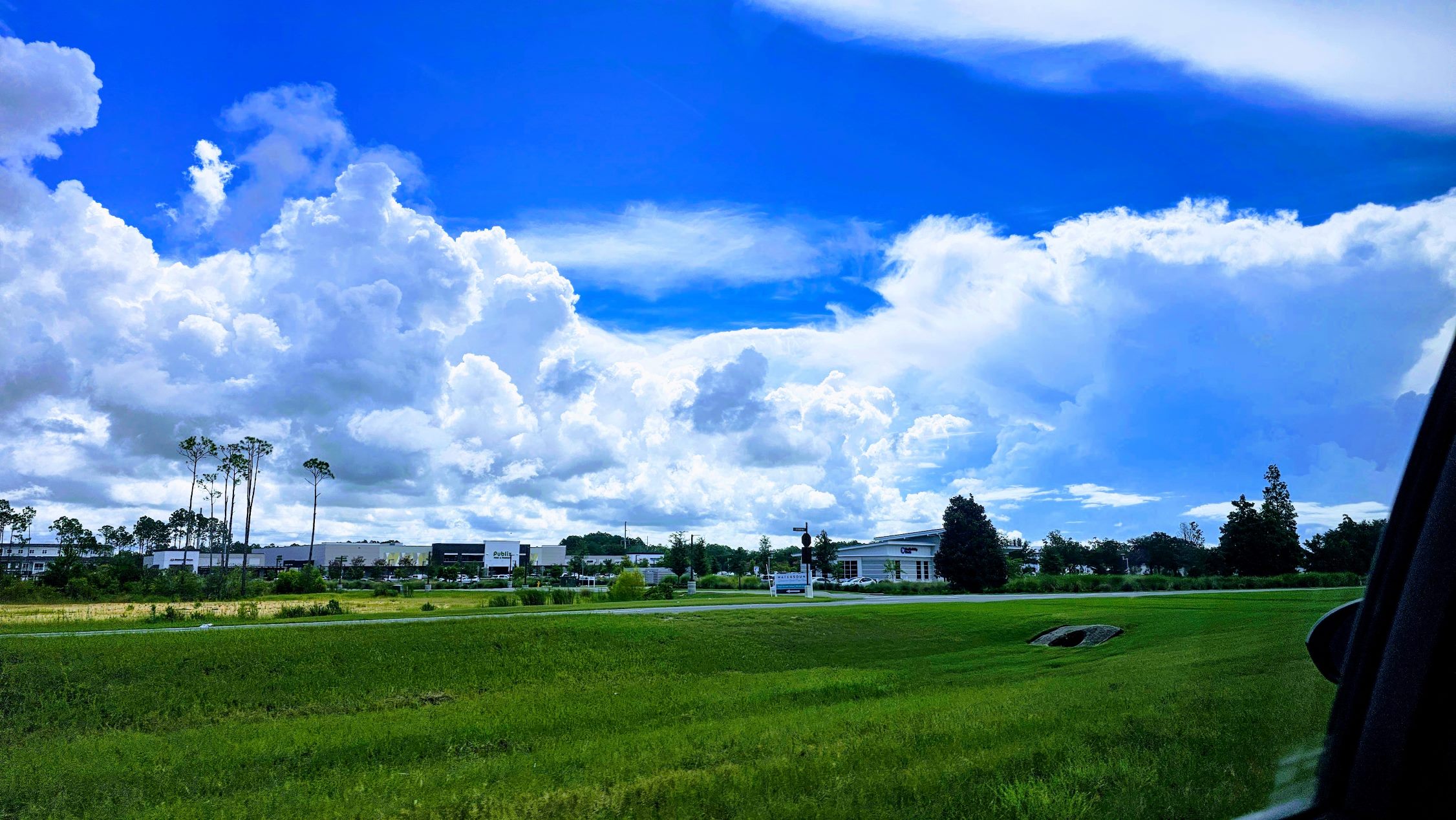 Vibrant blue sky with large, billowing white clouds over a lush green landscape, featuring commercial buildings in the distance and a car window frame visible on the right side.